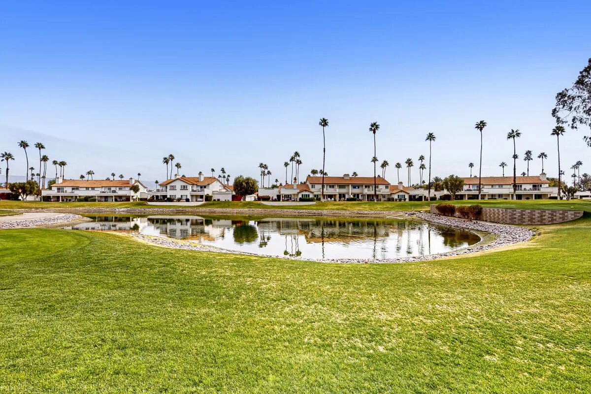 A landscaped pond sits amid green lawns, with low buildings and palm trees in the background.