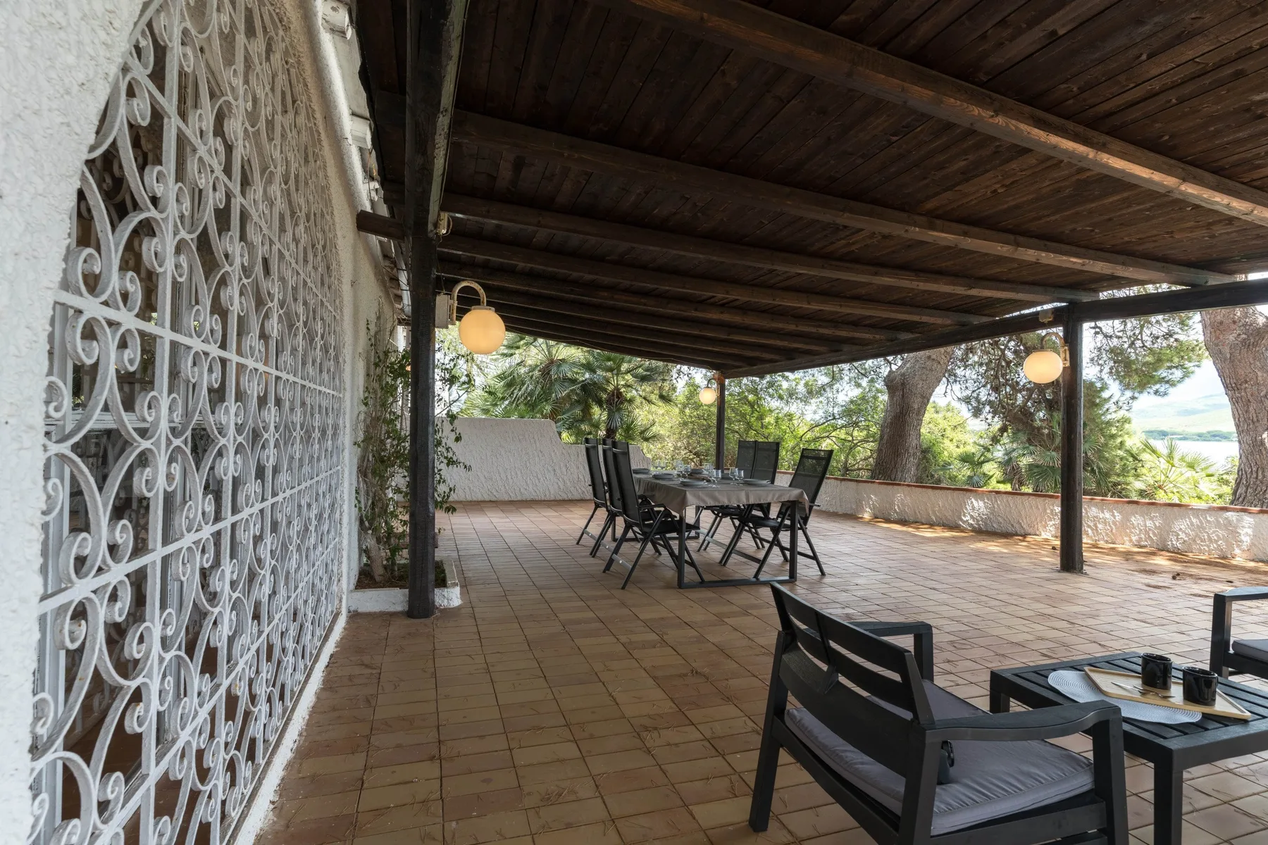 Una zona exterior cubierta para comer, con mesas y asientos, da a un paisaje frondoso.