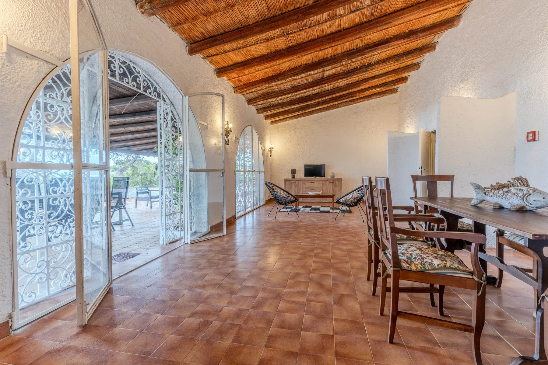 Zona de comedor y estar con mesas de madera, sillas y puertas acristaladas en arco.