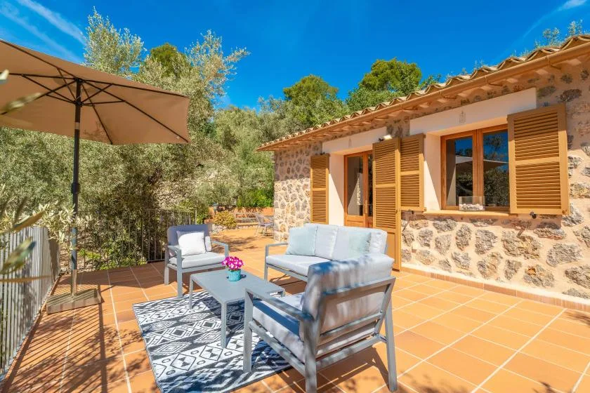 Un patio soleado junto a una casa de piedra tiene asientos, una mesa y una sombrilla grande.