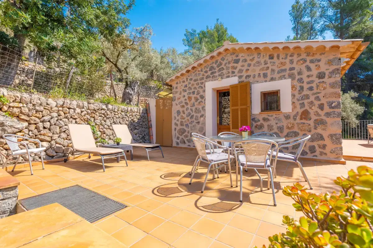 Un patio soleado junto a una casa de piedra, con asientos y tumbonas.