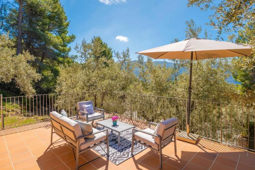 An outdoor patio with seating, a table, and a large umbrella overlooks leafy trees.