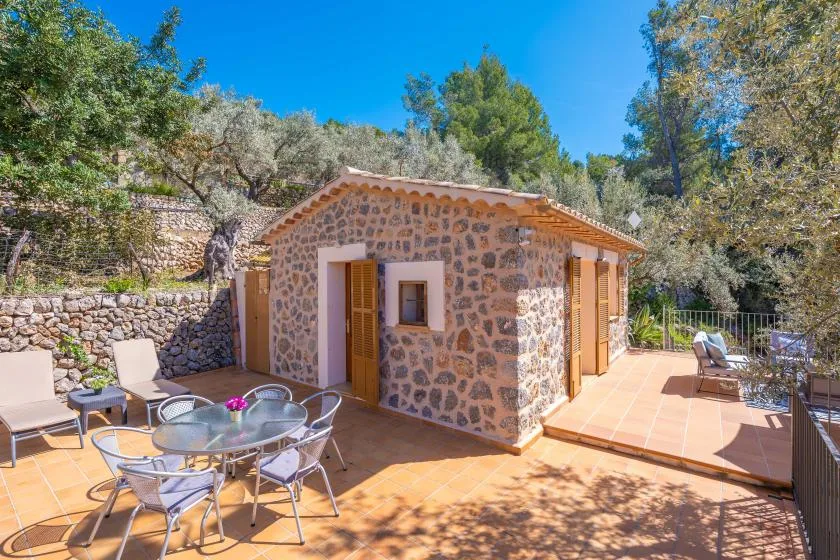 A stone cottage stands beside a sunny tiled patio with outdoor seating.
