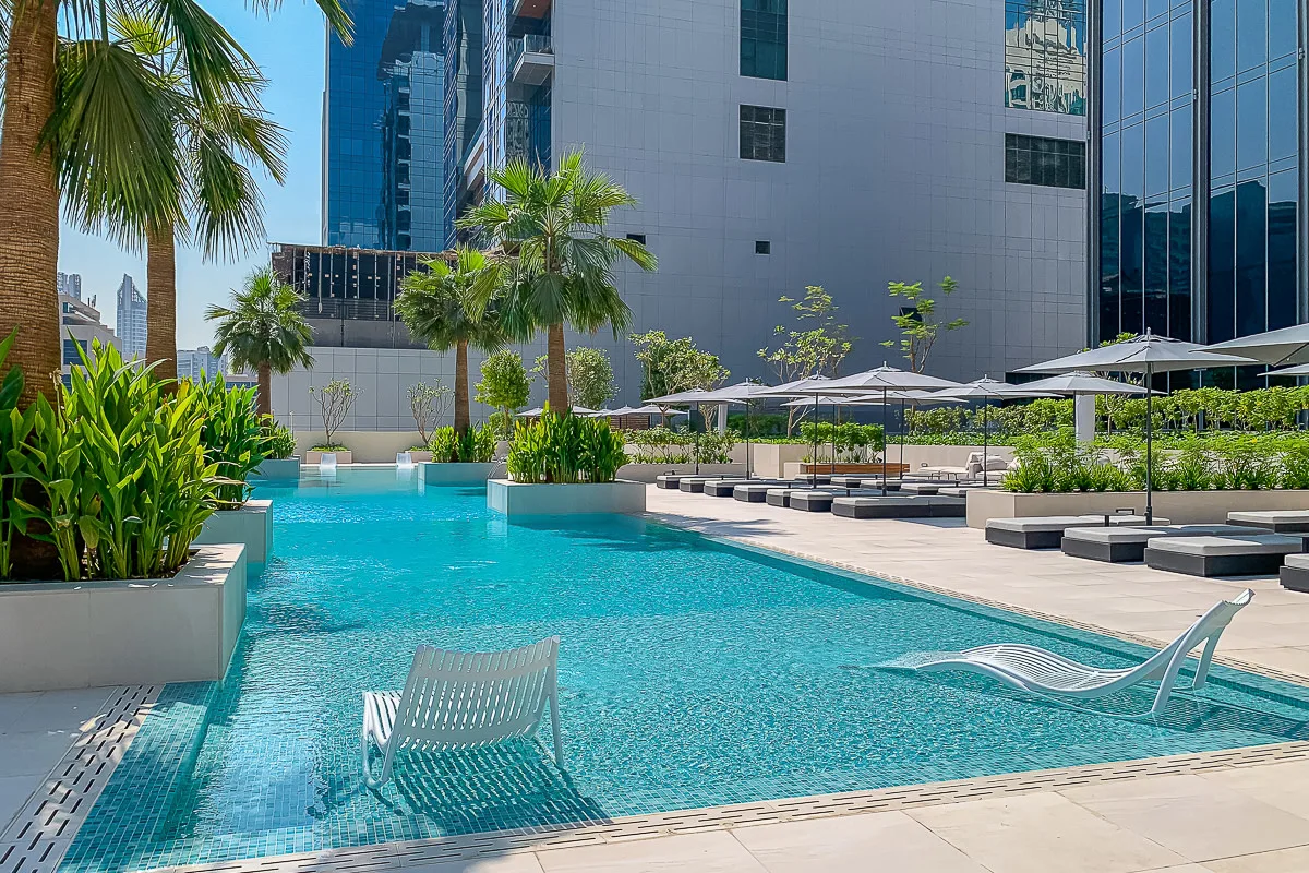 An outdoor pool beside modern buildings, with palm trees and lounge chairs.