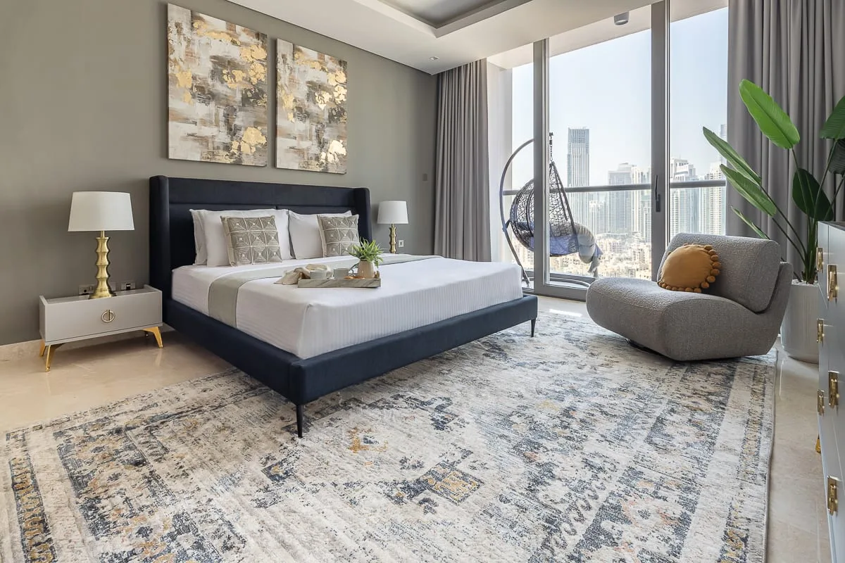 A bright bedroom with a large bed, patterned rug, and floor-to-ceiling windows.