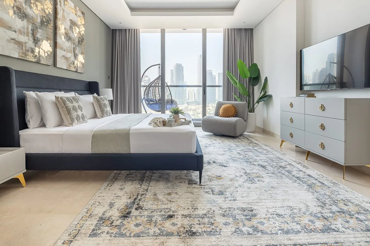 A modern bedroom with a large bed, patterned rug, and floor-to-ceiling windows.