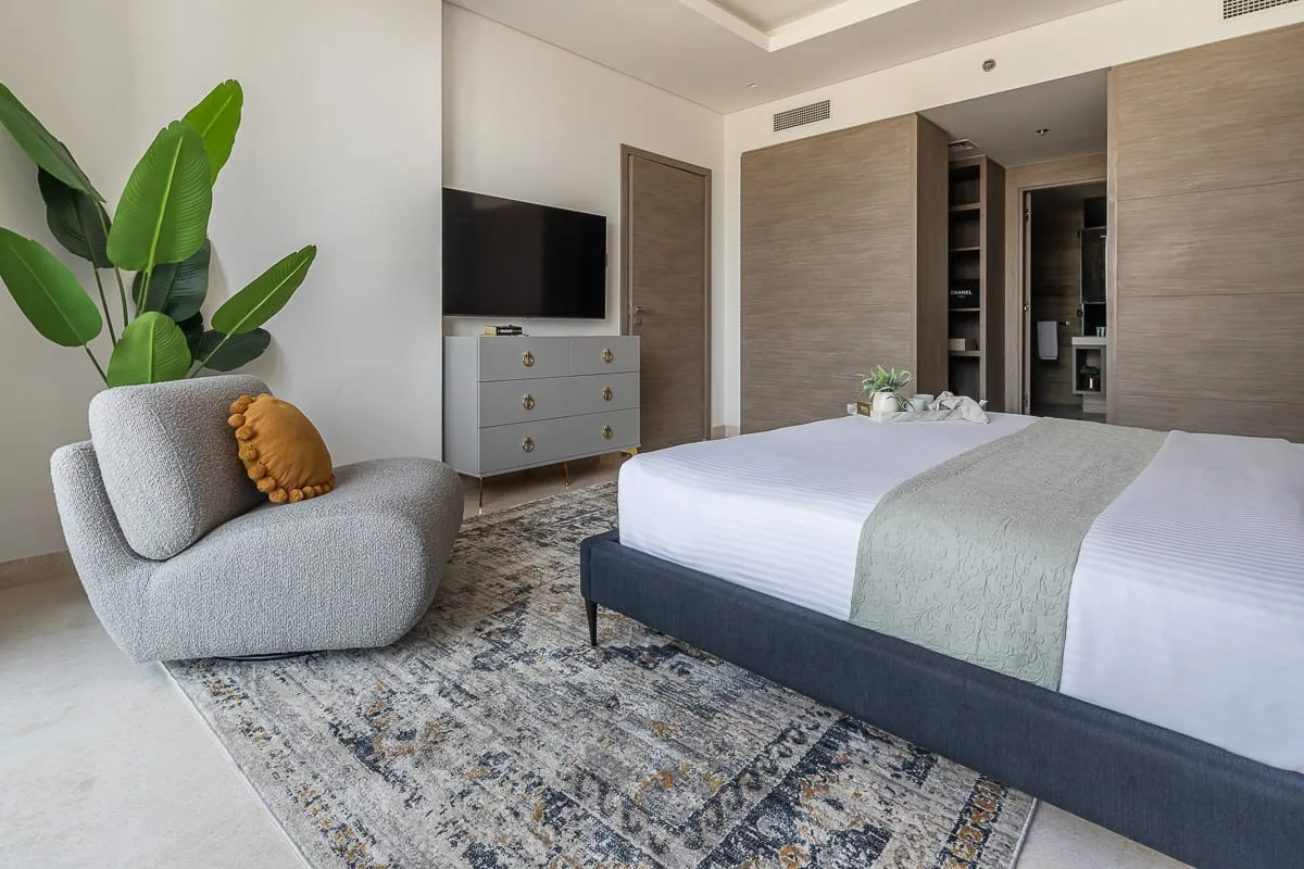 A bedroom with a neatly made bed, a lounge chair, and a television.