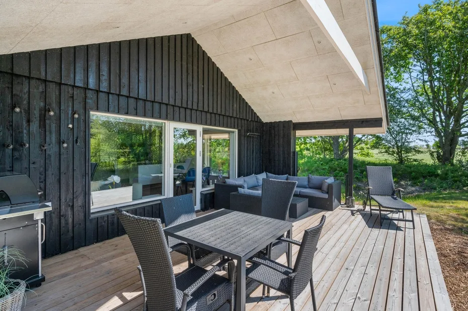 Una zona exterior de comedor y descanso está en una terraza de madera junto a grandes ventanas.