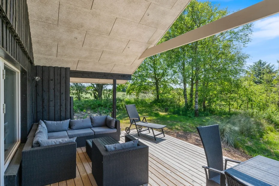 Una terraza exterior tiene asientos acolchados y sillas reclinables junto a una zona arbolada.
