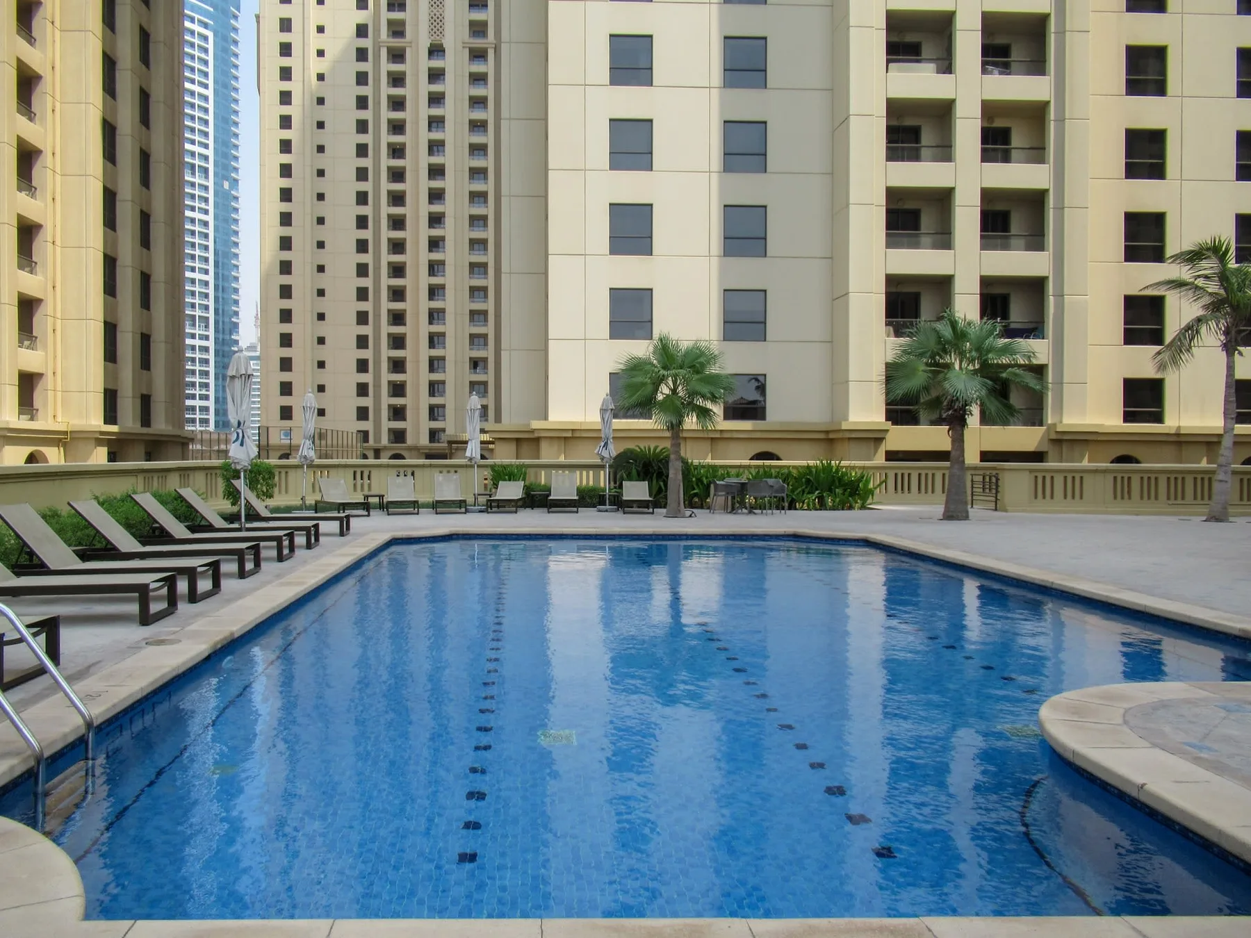 An outdoor swimming pool sits among tall apartment buildings and palm trees.