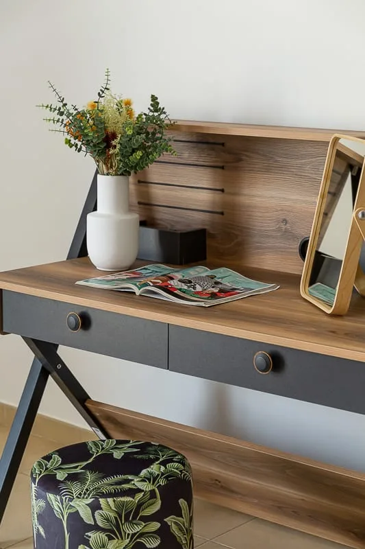 A wooden desk with a vase of flowers, an open magazine, and a patterned stool.