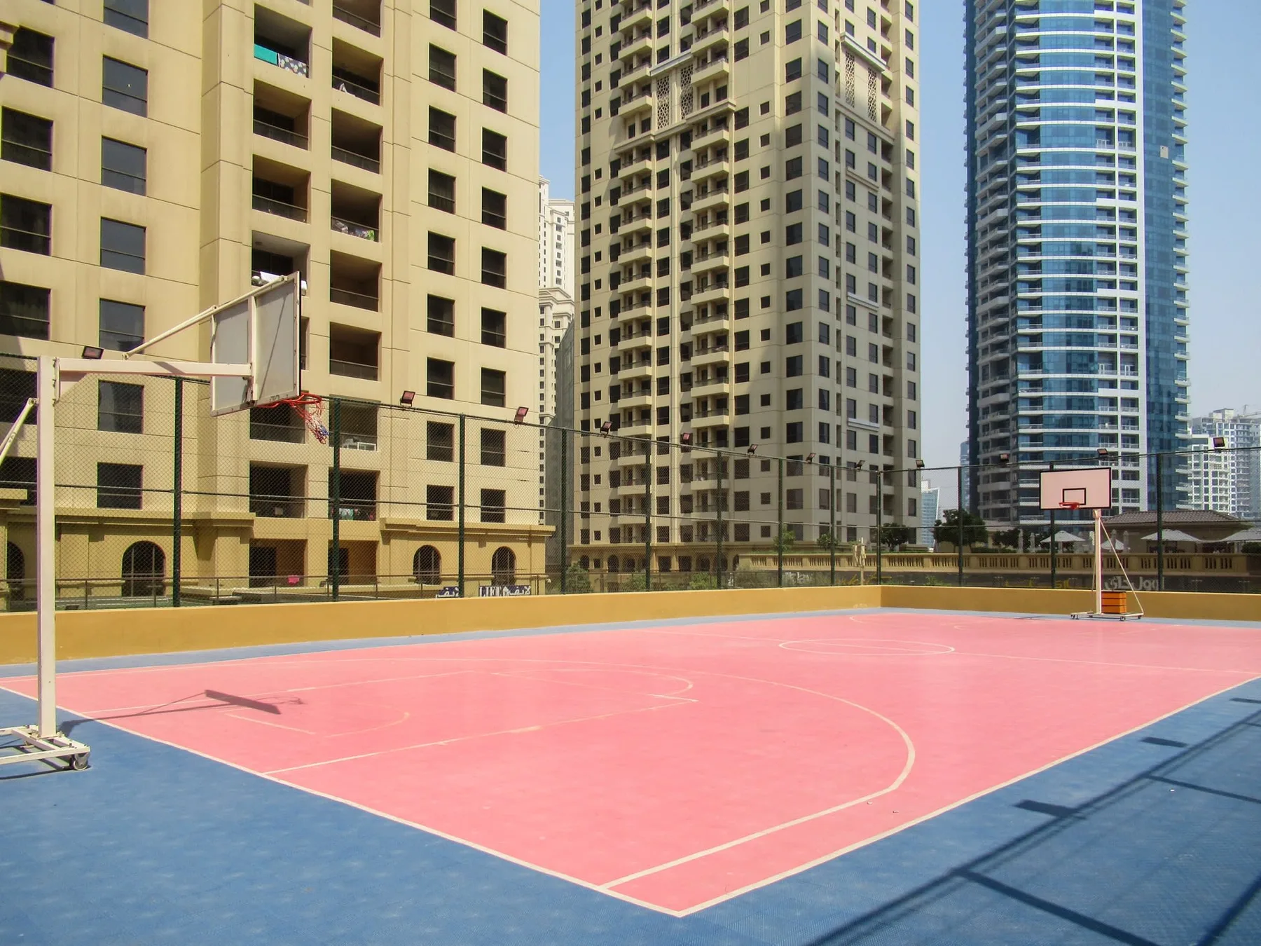 An outdoor basketball court with a pink playing surface and tall buildings in the background.