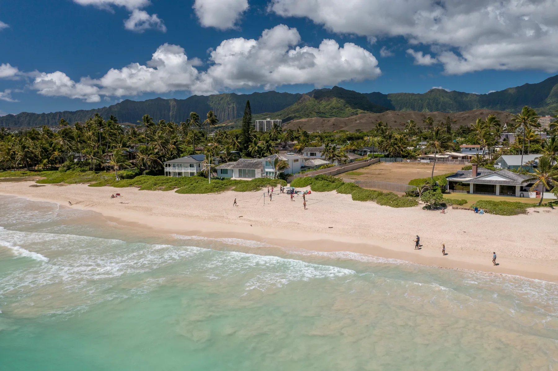 Una playa de arena bordea aguas turquesas poco profundas, con casas y montañas al fondo.