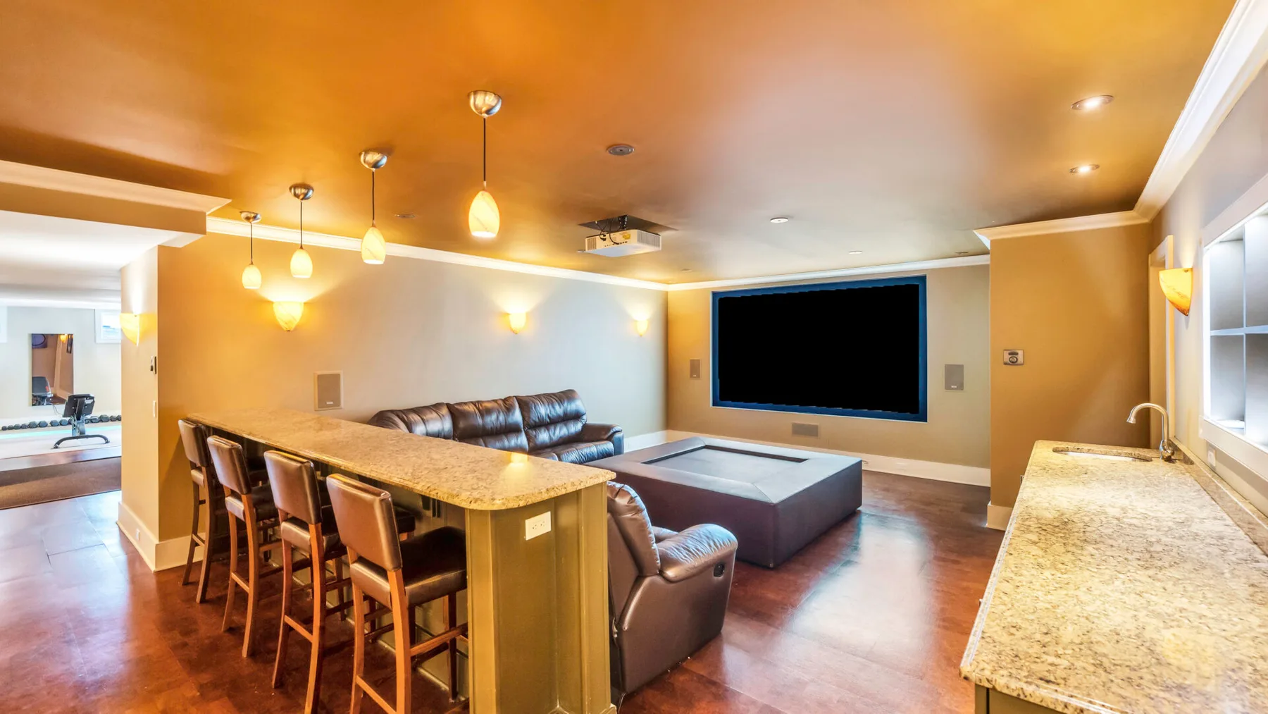Una sala de tonos cálidos cuenta con televisor, asientos y una isla de cocina con taburetes.
