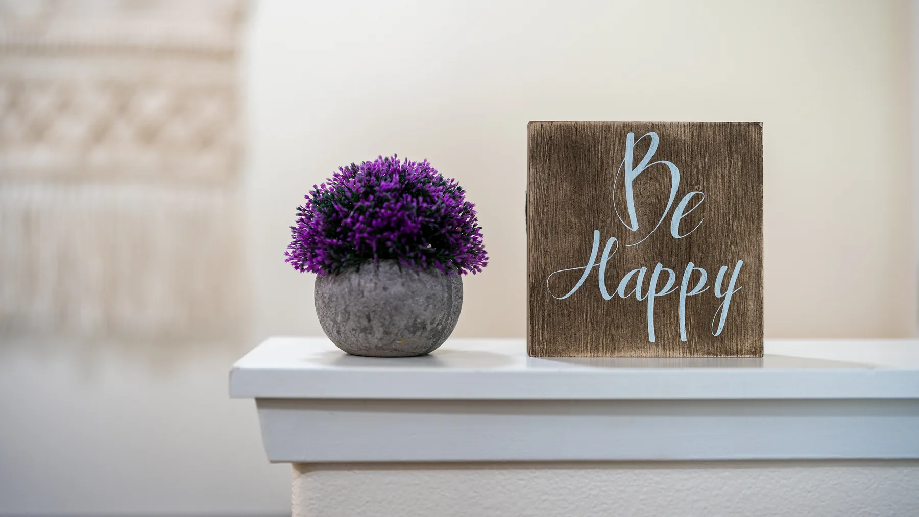 A small potted purple flower sits beside a wooden “Be Happy” sign on a white ledge.