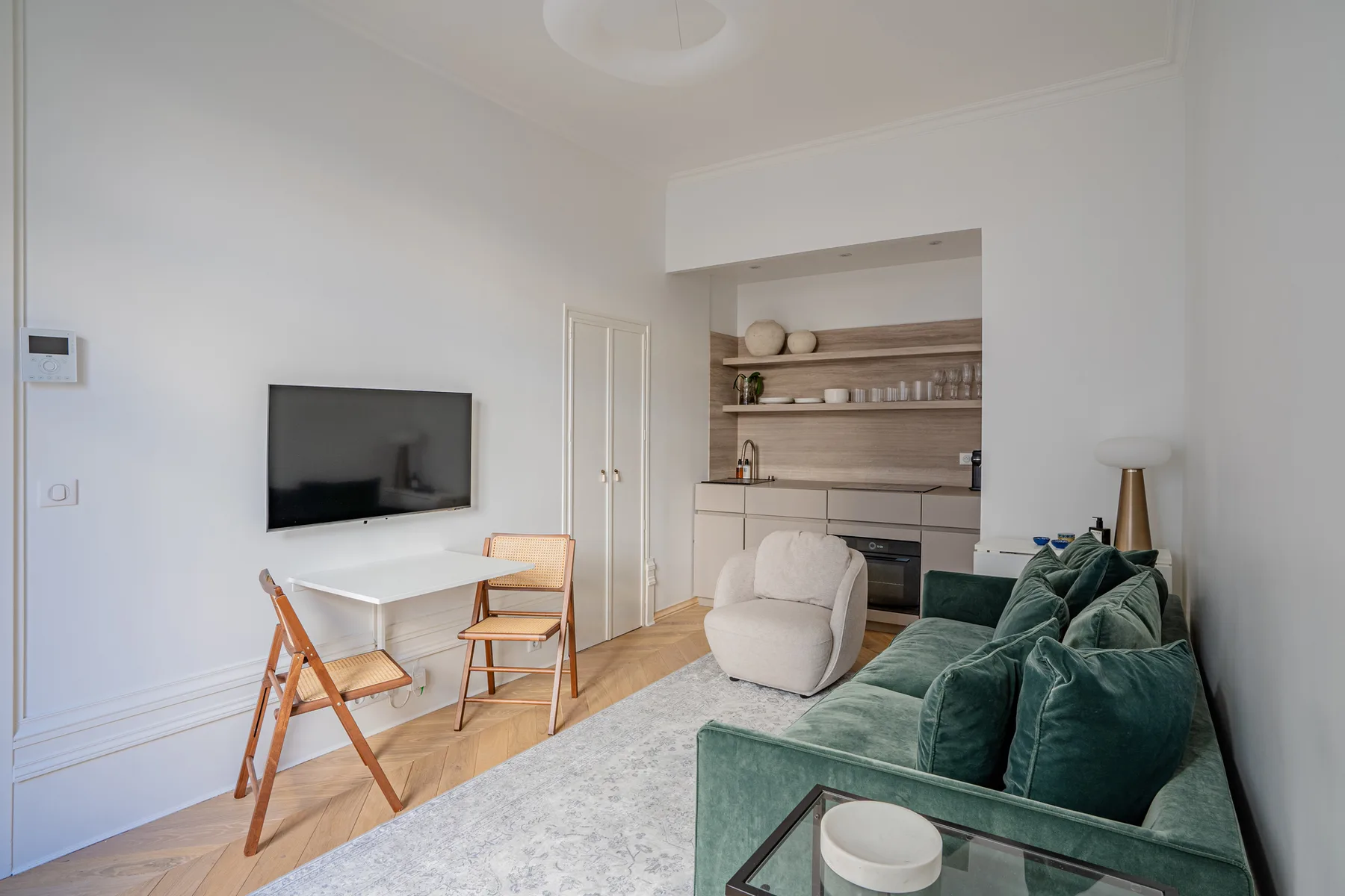 A bright living room with green seating, a television, and a compact kitchen.