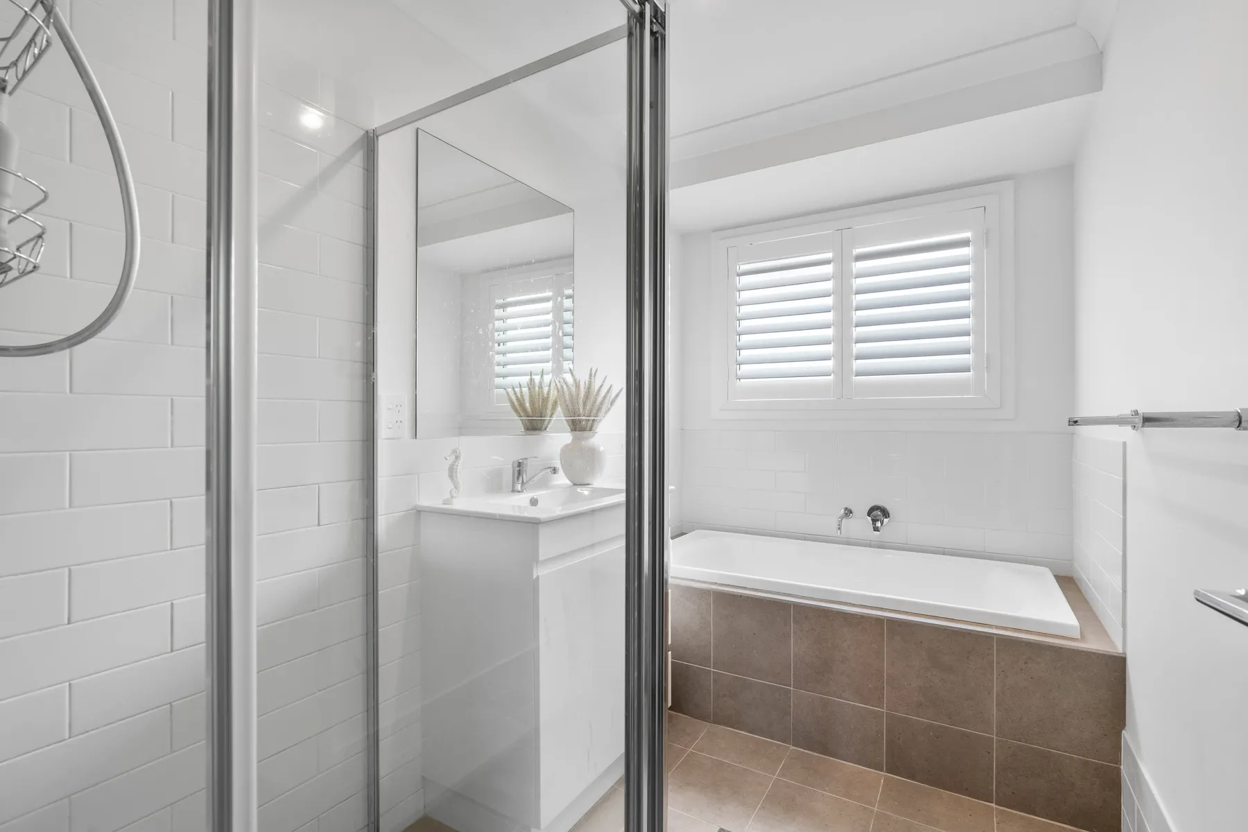 Un baño luminoso tiene una bañera bajo ventanas con contraventanas y una ducha de vidrio.