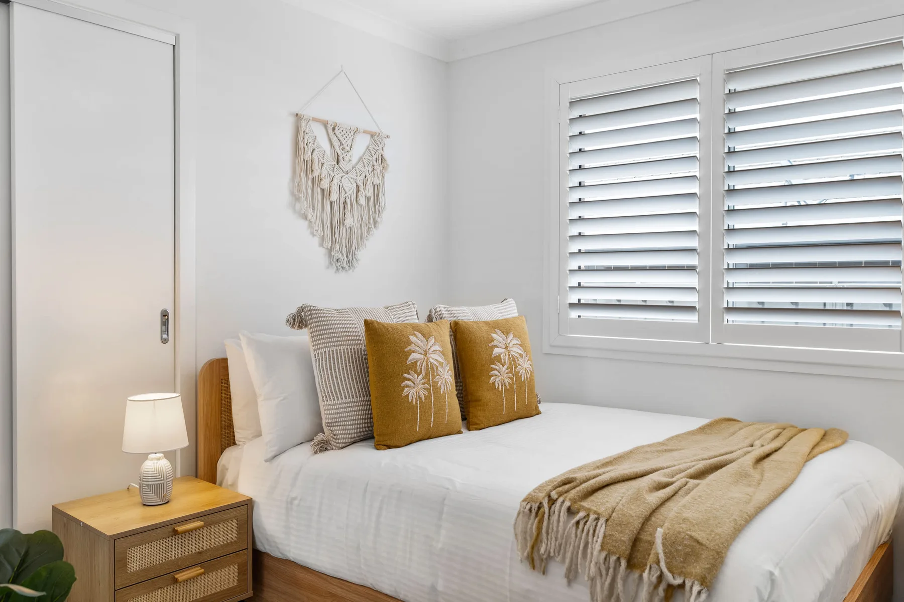 Un dormitorio luminoso con una cama arreglada y ventanas con persianas blancas.
