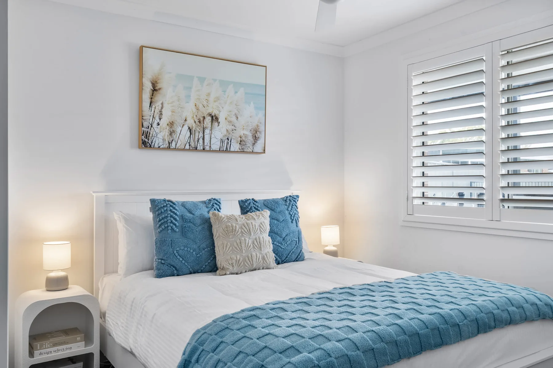Un dormitorio luminoso con una cama blanca, almohadas azules y ventanas amplias.