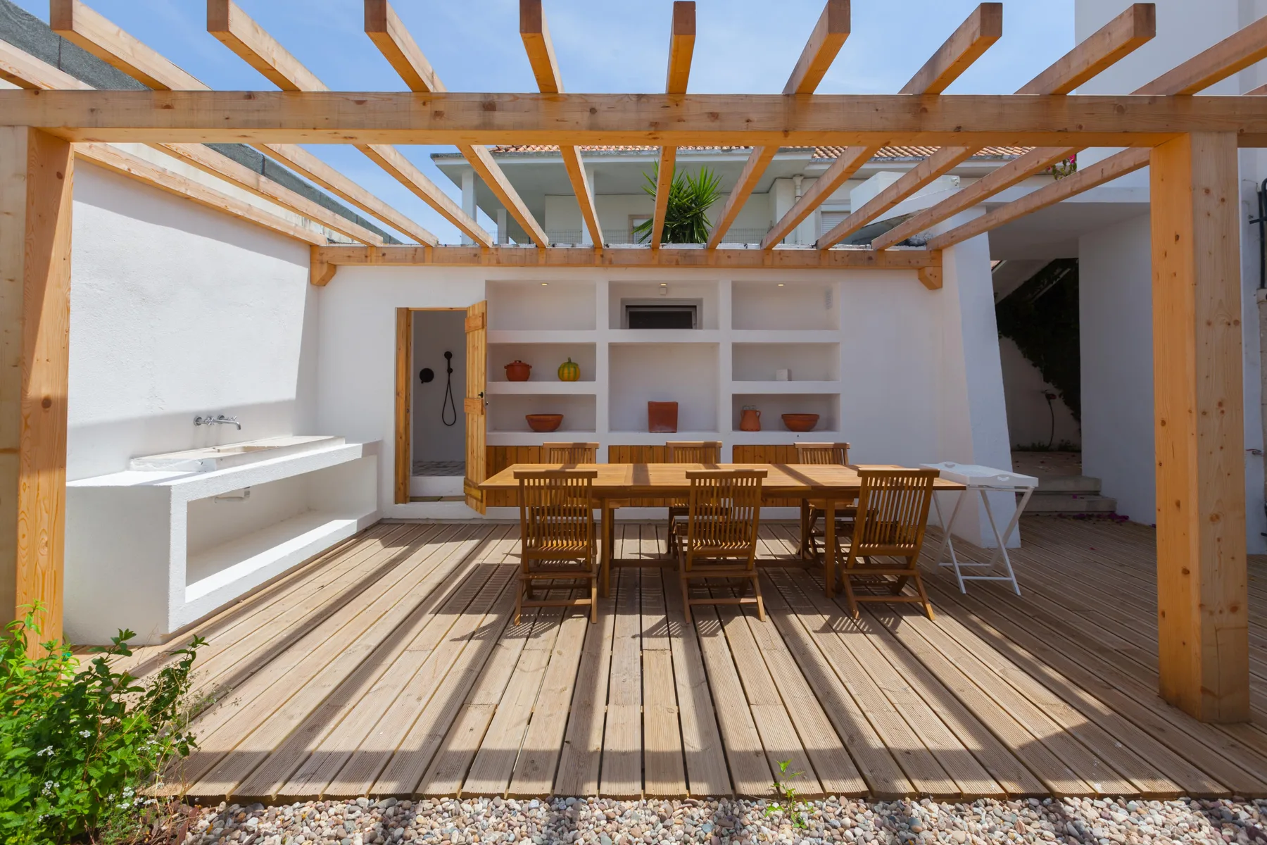 Una zona de comedor al aire libre está bajo una pérgola de madera, junto a una encimera y estantes abiertos.