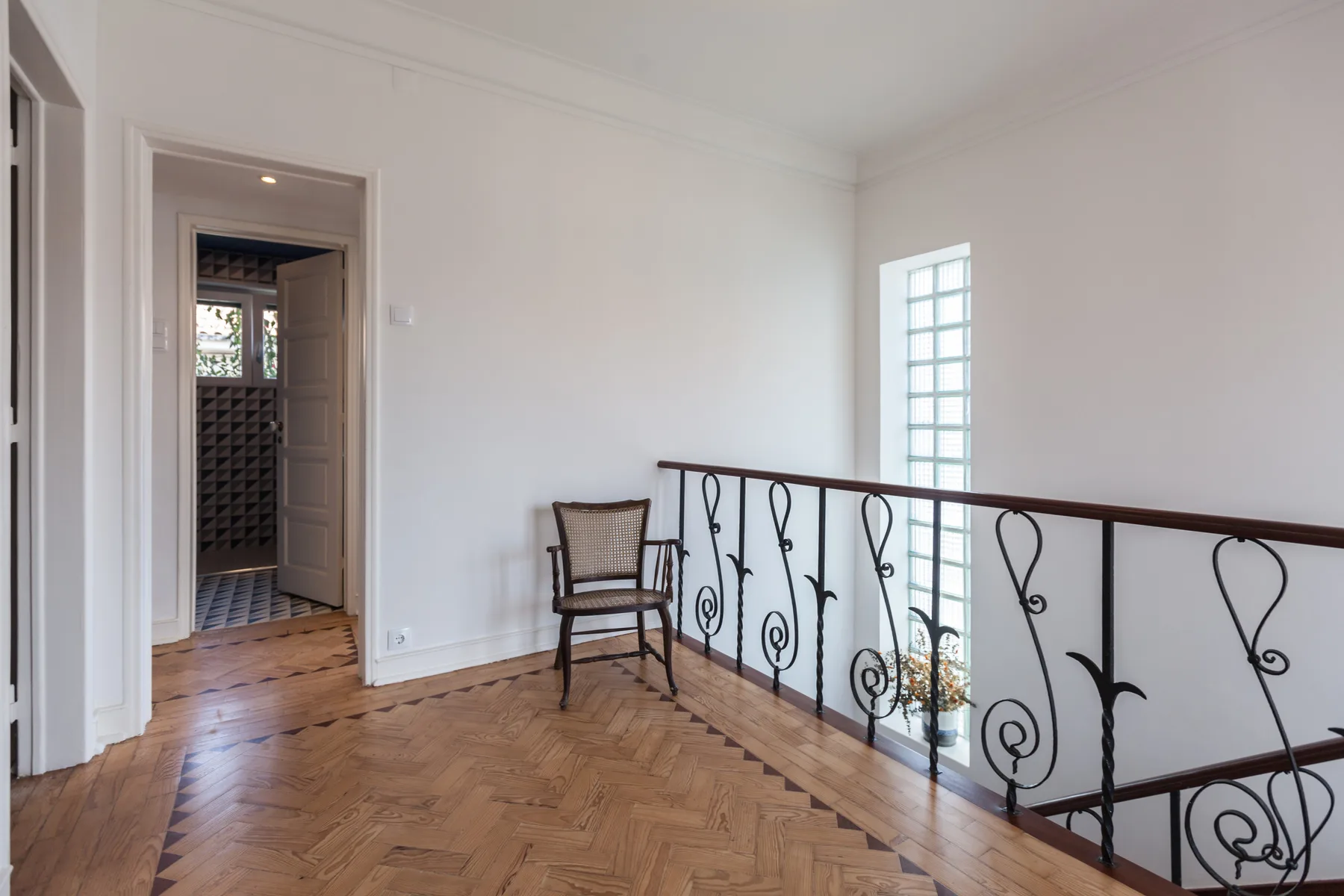Un descansillo luminoso con suelo de parquet, una silla y una barandilla decorativa de metal.