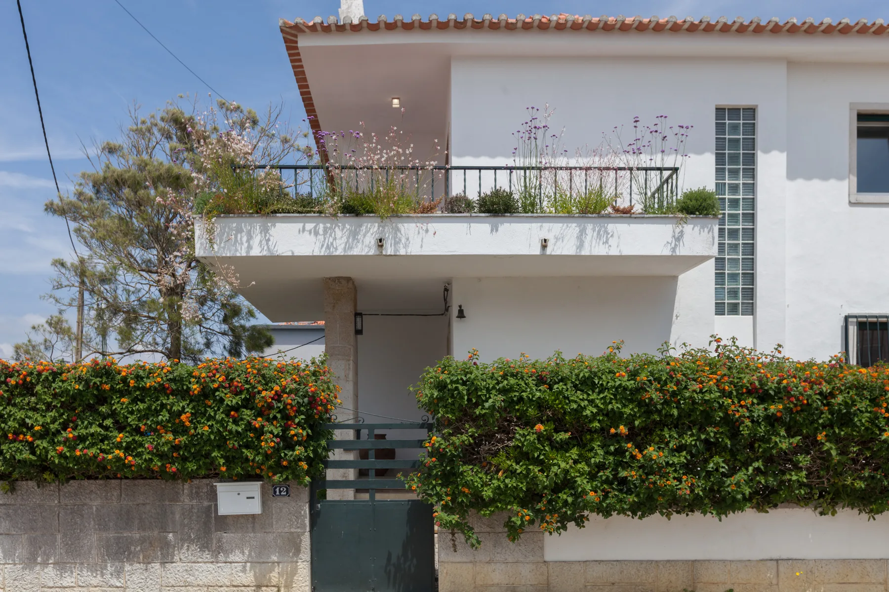 Exterior de una casa blanca con balcón y arbustos floridos junto al muro frontal.