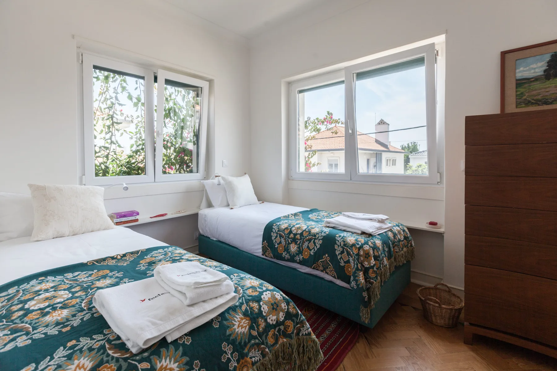 Un dormitorio luminoso con dos camas, ropa de cama estampada y ventanas grandes.