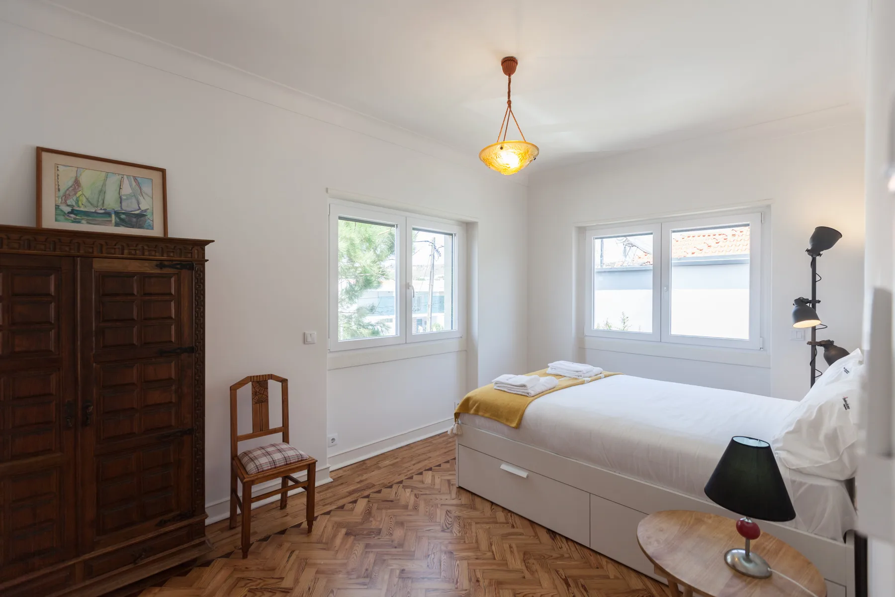 Un dormitorio luminoso con una cama junto a dos ventanas y suelo de madera.