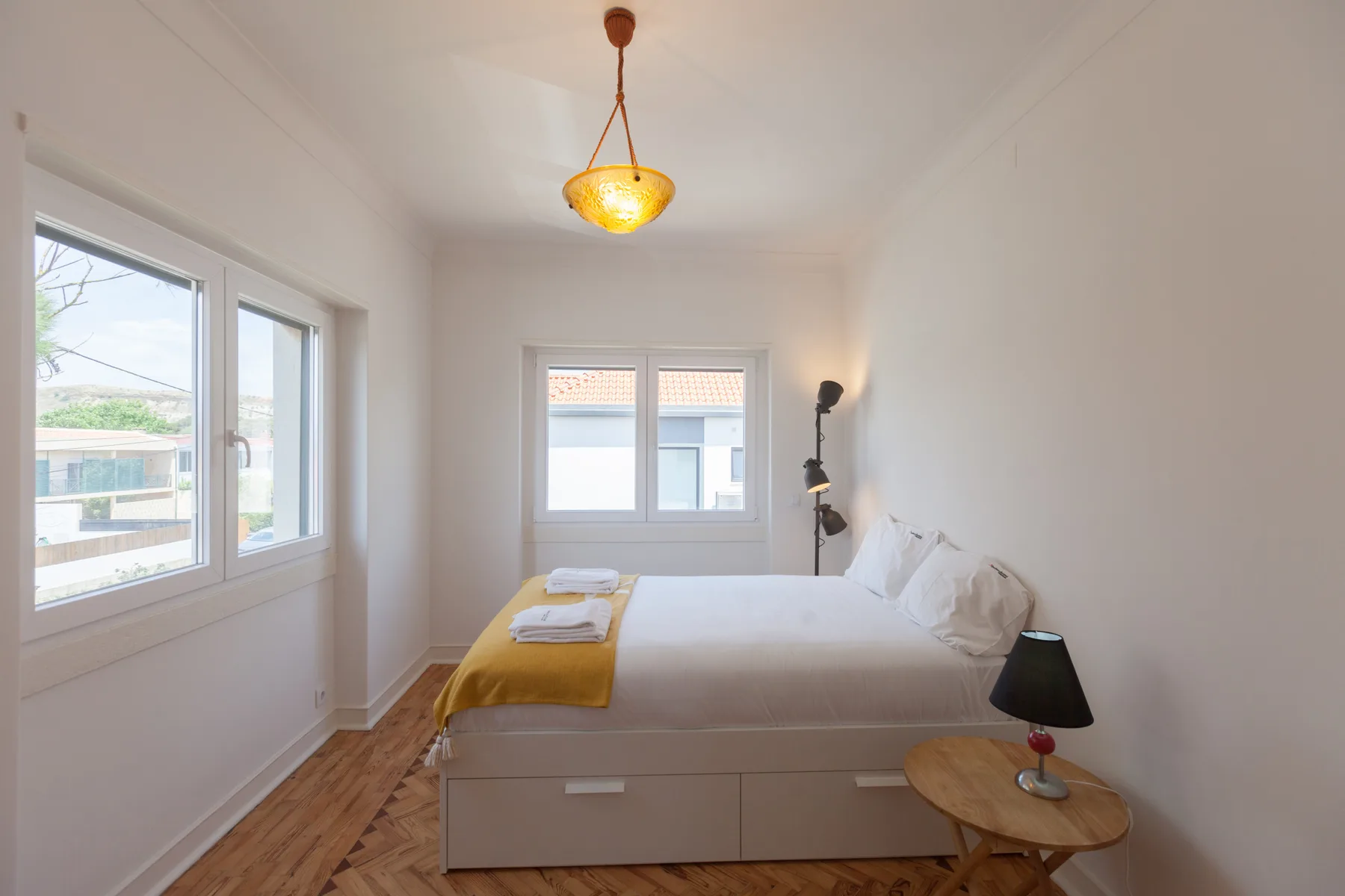 Un dormitorio luminoso con una cama, una colcha mostaza y ventanas.