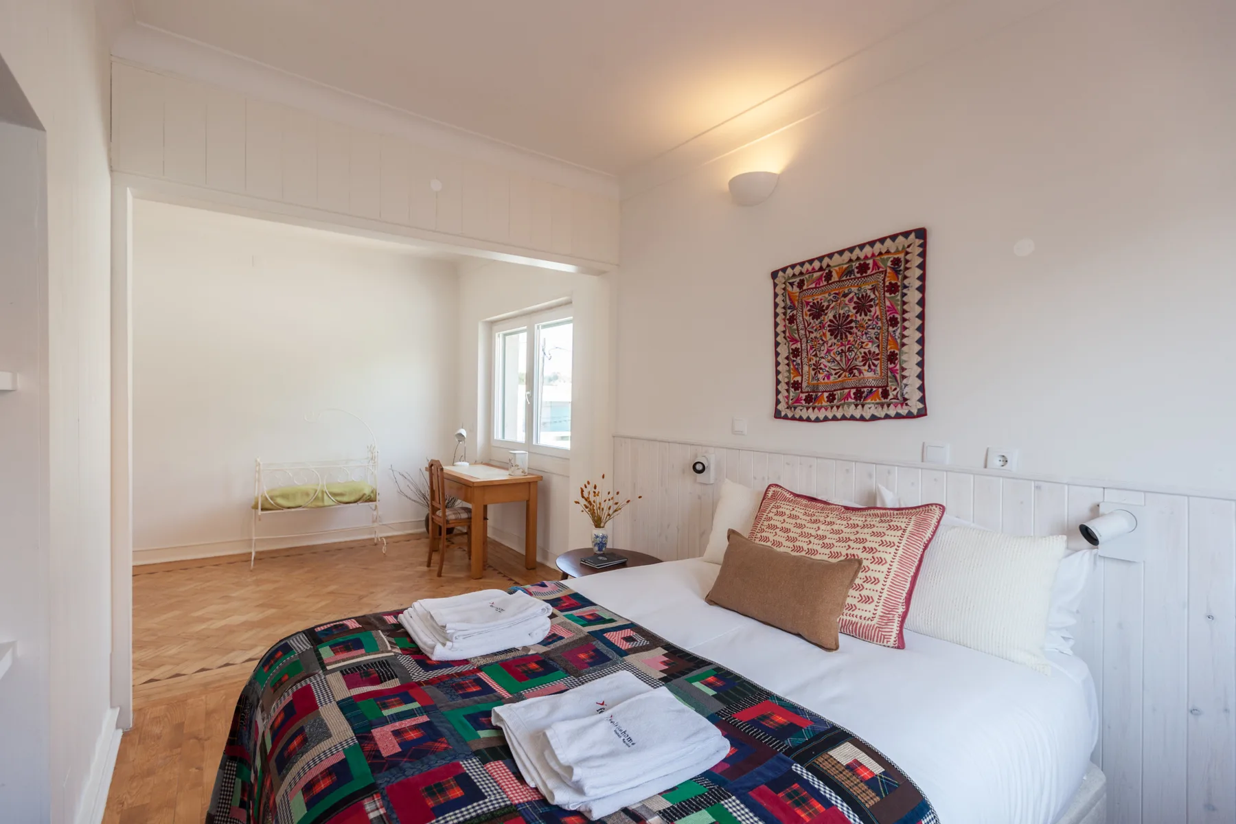 Un dormitorio con una cama cubierta por una manta colorida estampada y un pequeño escritorio junto a la ventana.