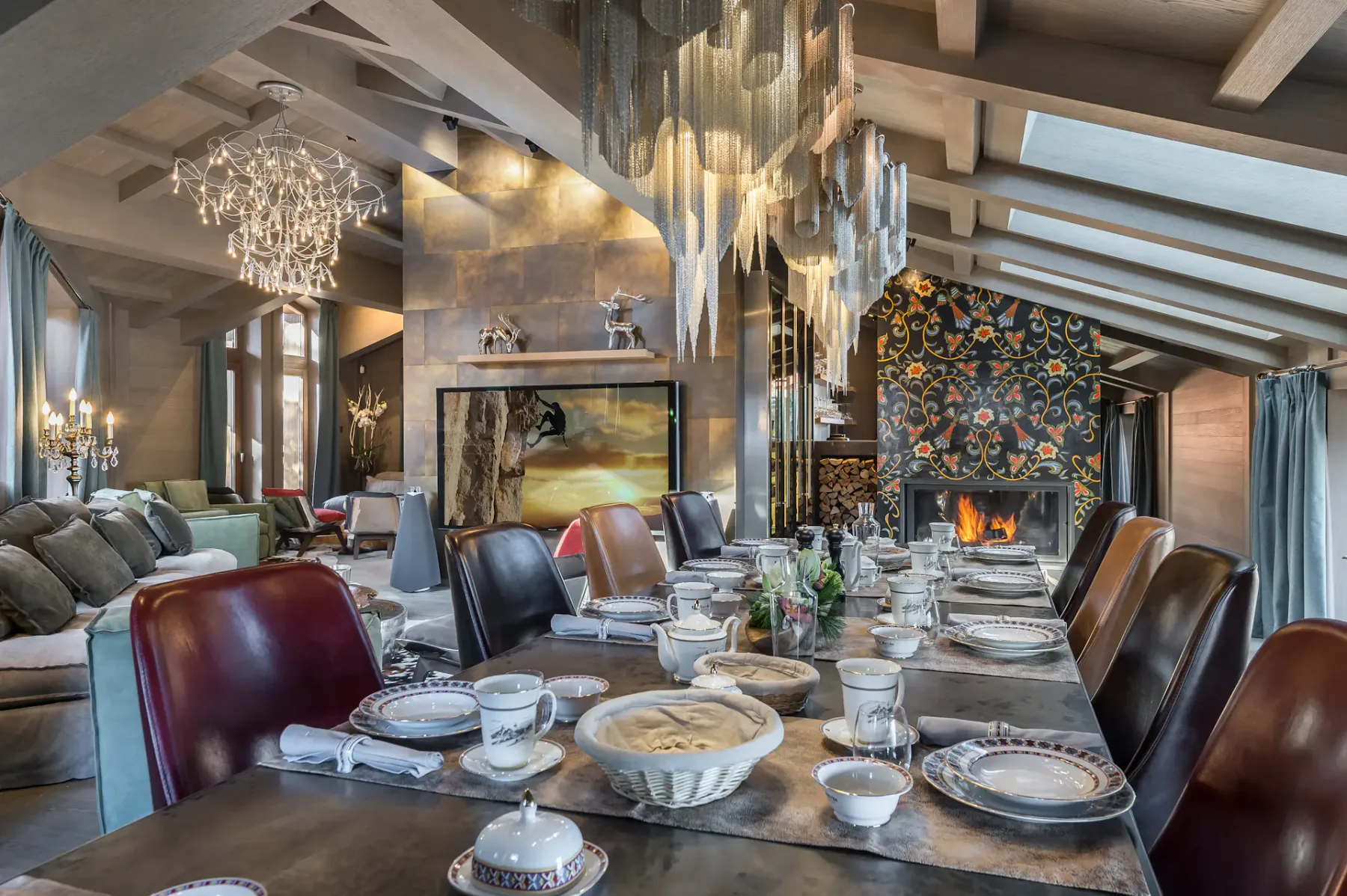 Luxurious dining room with a long table set for a meal, elegant chandeliers, a fireplace, and large windows with curtains.