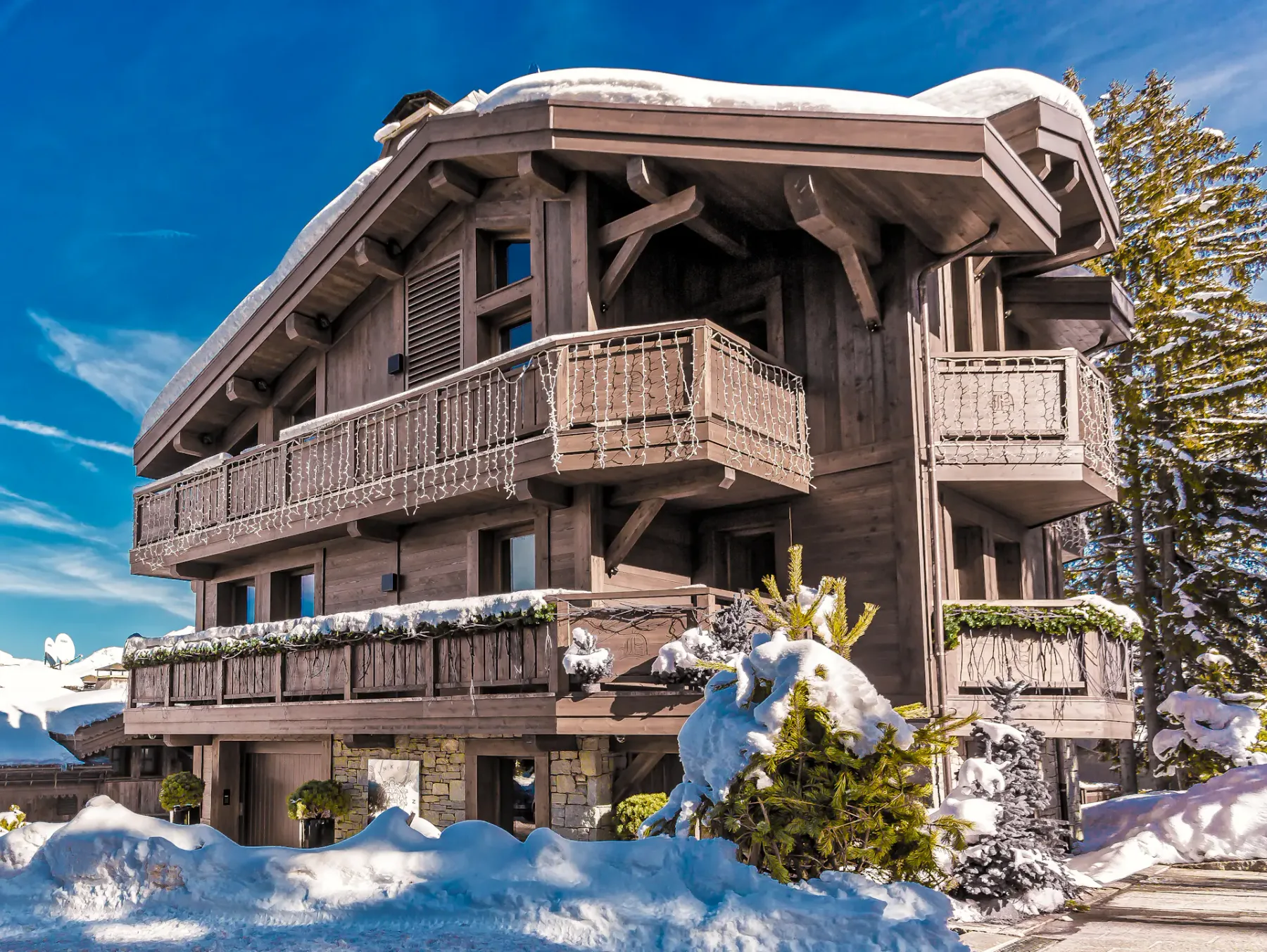 Chalet de madera con el tejado y los balcones cubiertos de nieve, rodeado de nieve y árboles de hoja perenne, bajo un cielo azul despejado.