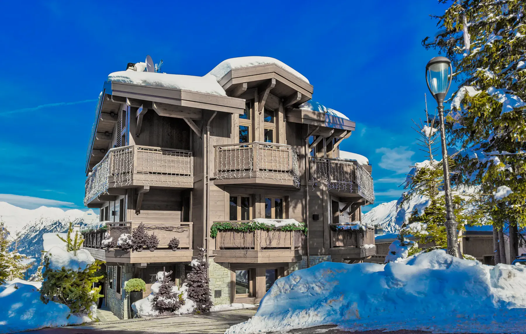 Chalet cubierto de nieve con balcones de madera, rodeado de árboles cargados de nieve y montañas bajo un cielo azul despejado.