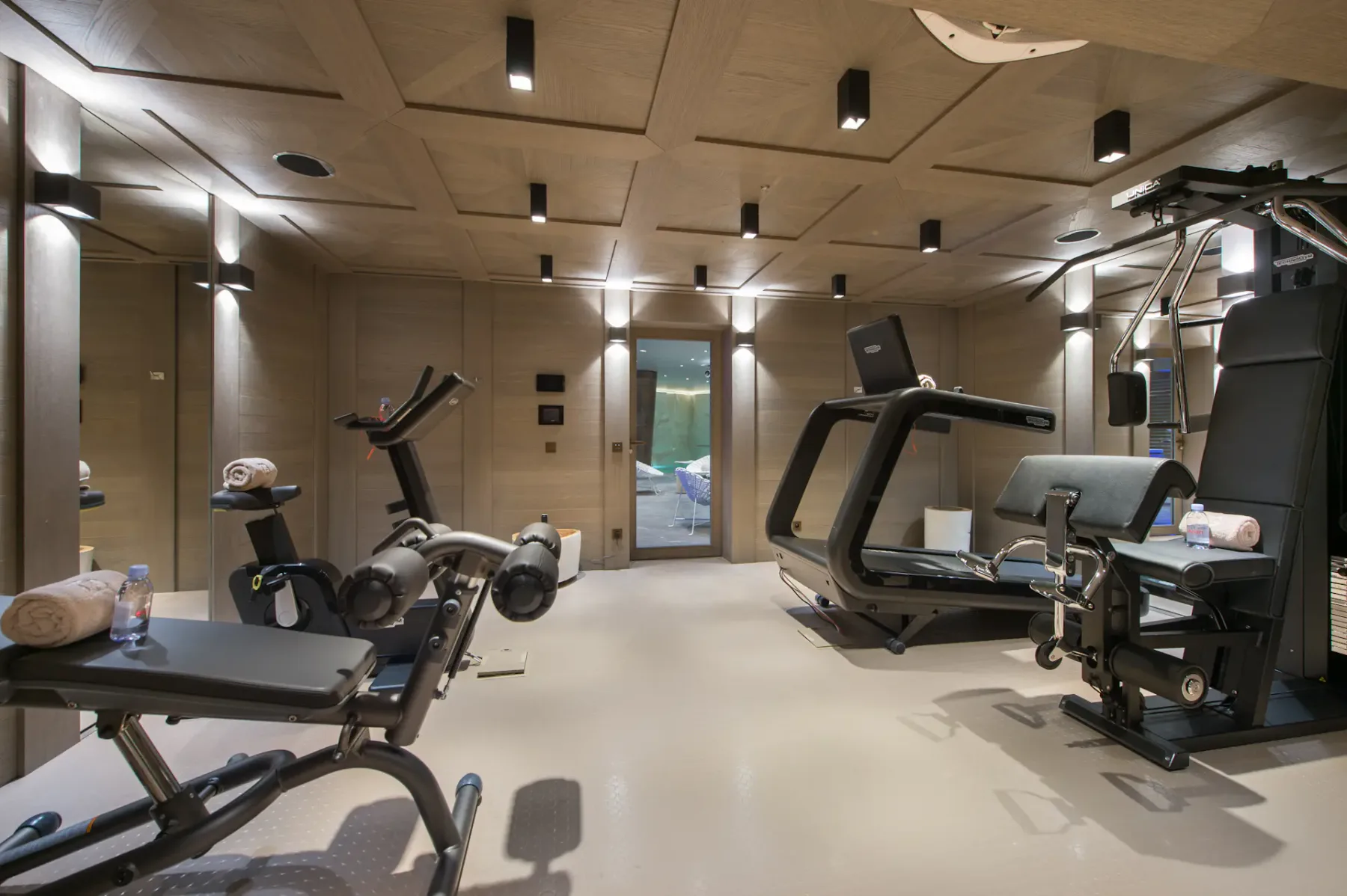 Gimnasio doméstico moderno con aparatos de ejercicio, entre ellos una cinta de correr, una bicicleta estática y un banco de pesas. Iluminación suave y paneles de madera en el techo.