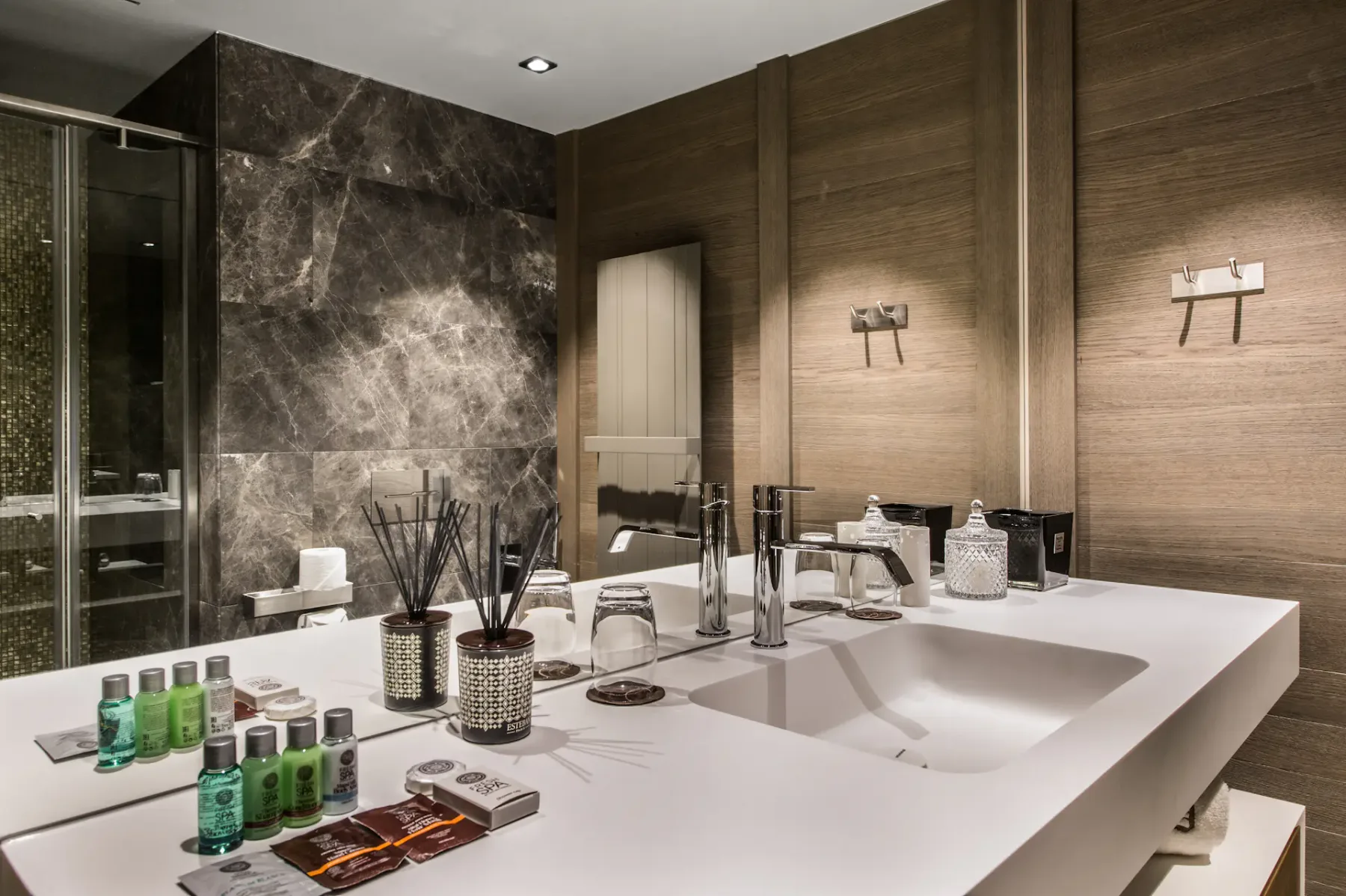 Baño moderno con una elegante encimera blanca, dos lavabos, artículos de aseo y un espejo grande. Los detalles de mármol oscuro y madera realzan la decoración.