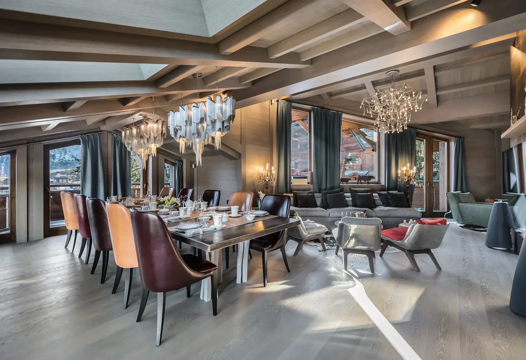 Amplia zona de comedor y estar con vigas de madera, elegantes lámparas de araña, una mesa grande, sillas de cuero y vistas panorámicas a las montañas a través de las ventanas.