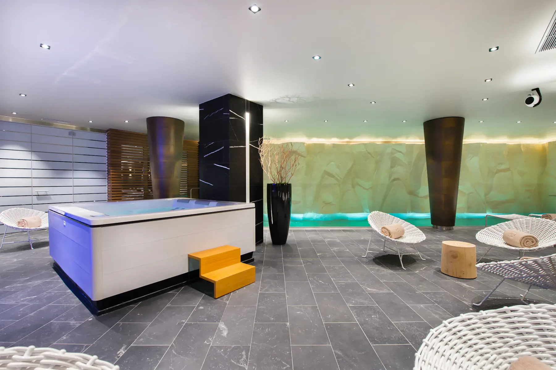 Moderna sala de spa con un jacuzzi blanco, escalones naranjas, sillones de mimbre e iluminación ambiental. El suelo de piedra y las paredes texturizadas realzan el ambiente sereno.