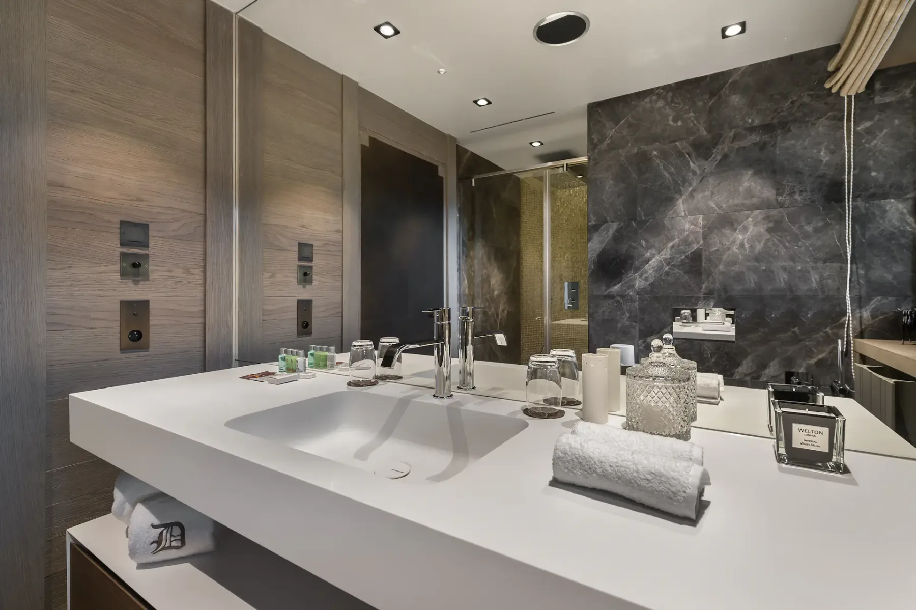 Baño moderno con una encimera blanca, un lavabo y un grifo; las toallas y los artículos de aseo están ordenados cuidadosamente. Los detalles de madera oscura y piedra realzan la decoración.