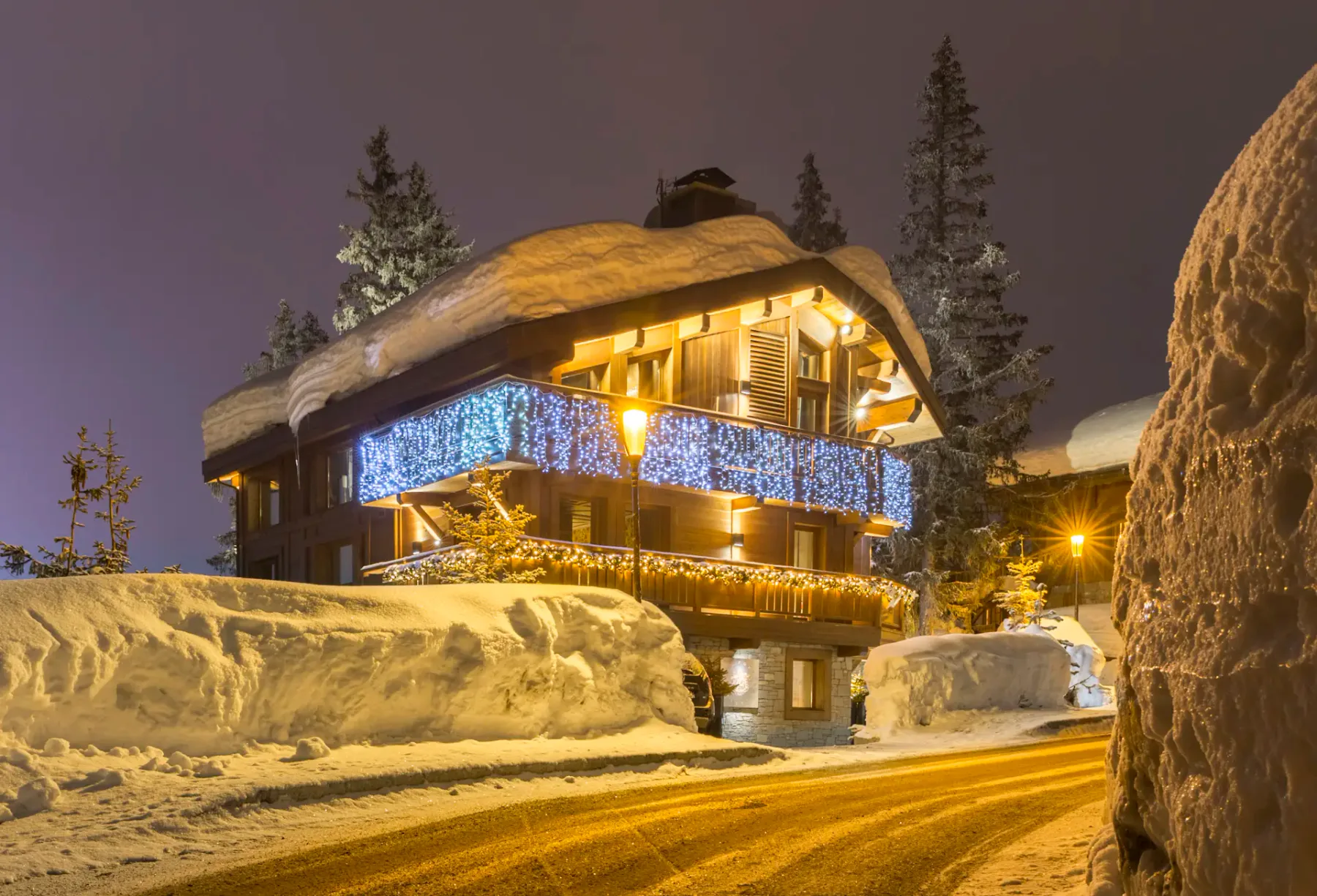 Chalet cubierto de nieve adornado con luces azules y blancas, rodeado de altos bancos de nieve y árboles, bajo un cielo violeta al anochecer.