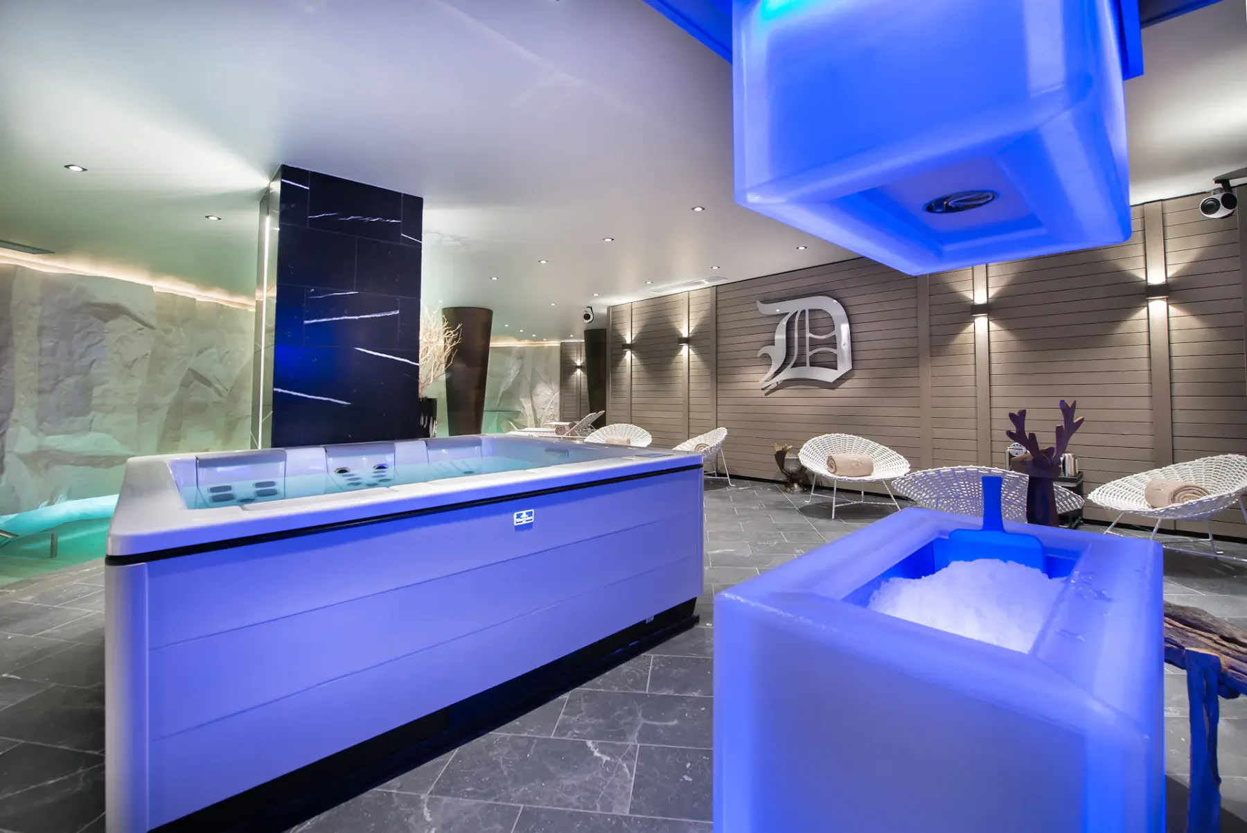 Moderna sala de spa con una bañera de hidromasaje iluminada en azul, un baño de hielo, tumbonas y un elegante diseño en la pared.