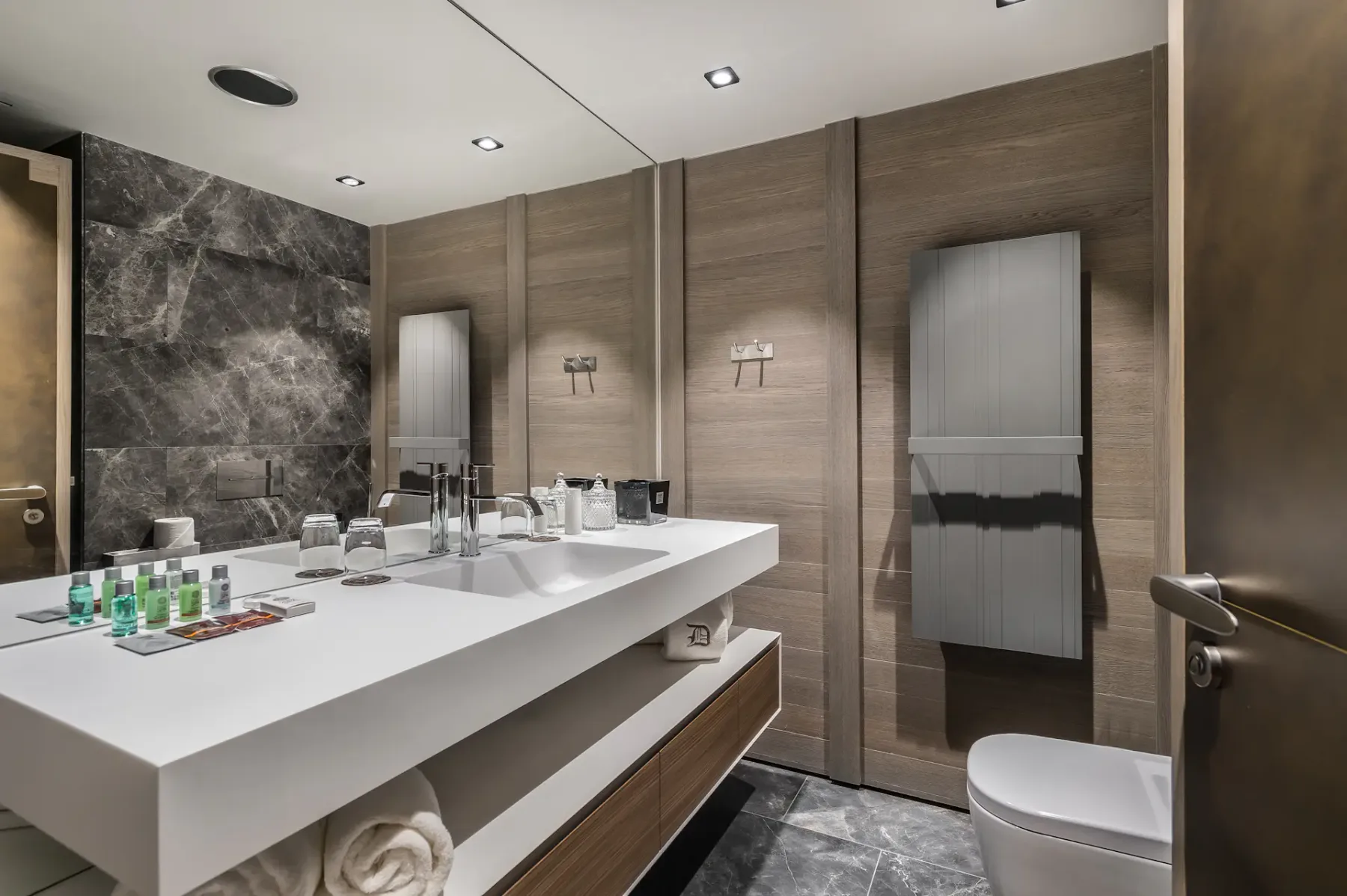 Baño moderno con una elegante encimera blanca, dos lavabos, artículos de aseo, detalles de madera, un espejo grande y revestimiento de mármol gris en las paredes.
