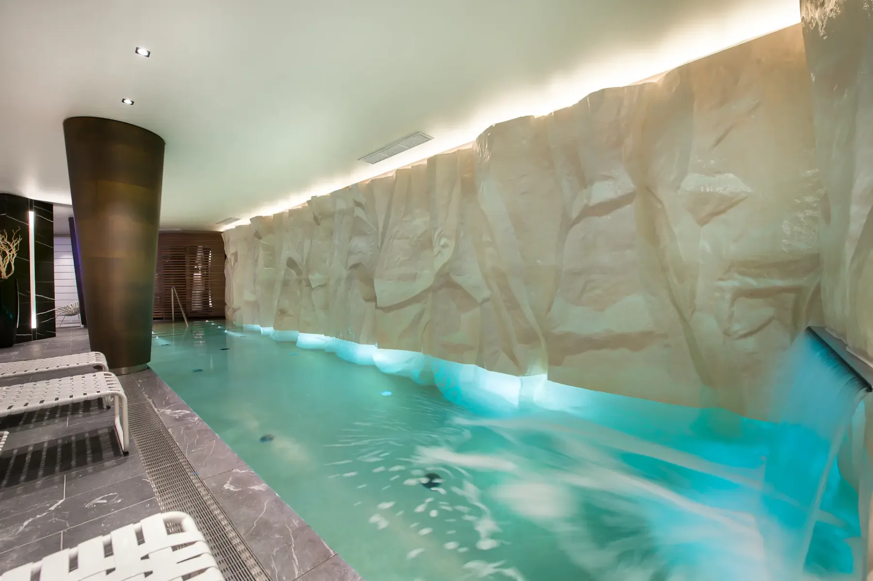 Piscina cubierta con agua turquesa, una cascada y tumbonas sobre un suelo de baldosas, bajo una suave iluminación de techo.