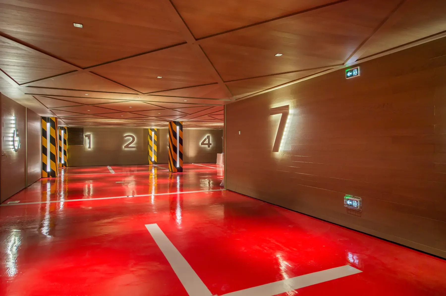 Garaje moderno con suelo rojo brillante, paredes de madera y grandes números iluminados: 1, 2, 4 y 7. Las columnas a rayas aportan contraste.