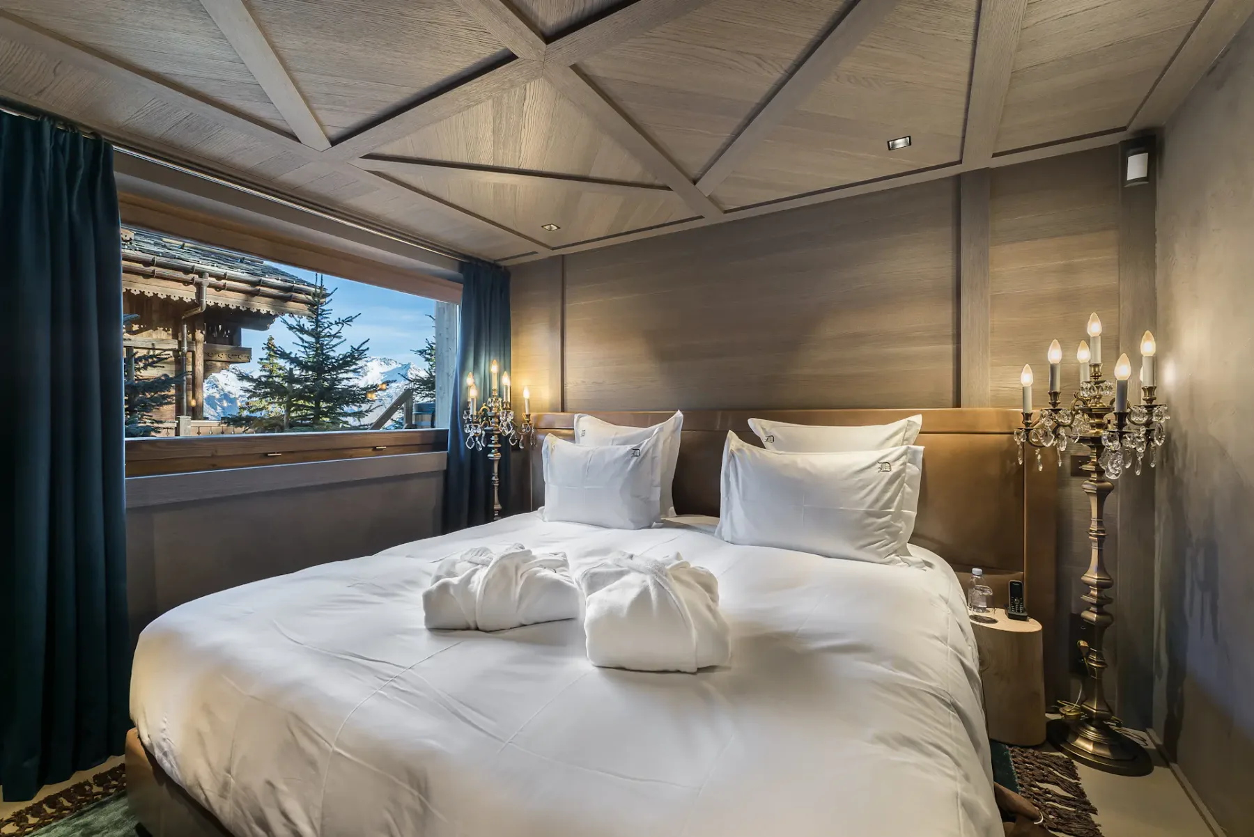 Acogedor dormitorio con una cama grande, ropa de cama blanca y candelabros decorativos. Una ventana ofrece vistas a las montañas nevadas y a una cabaña de madera en el exterior.