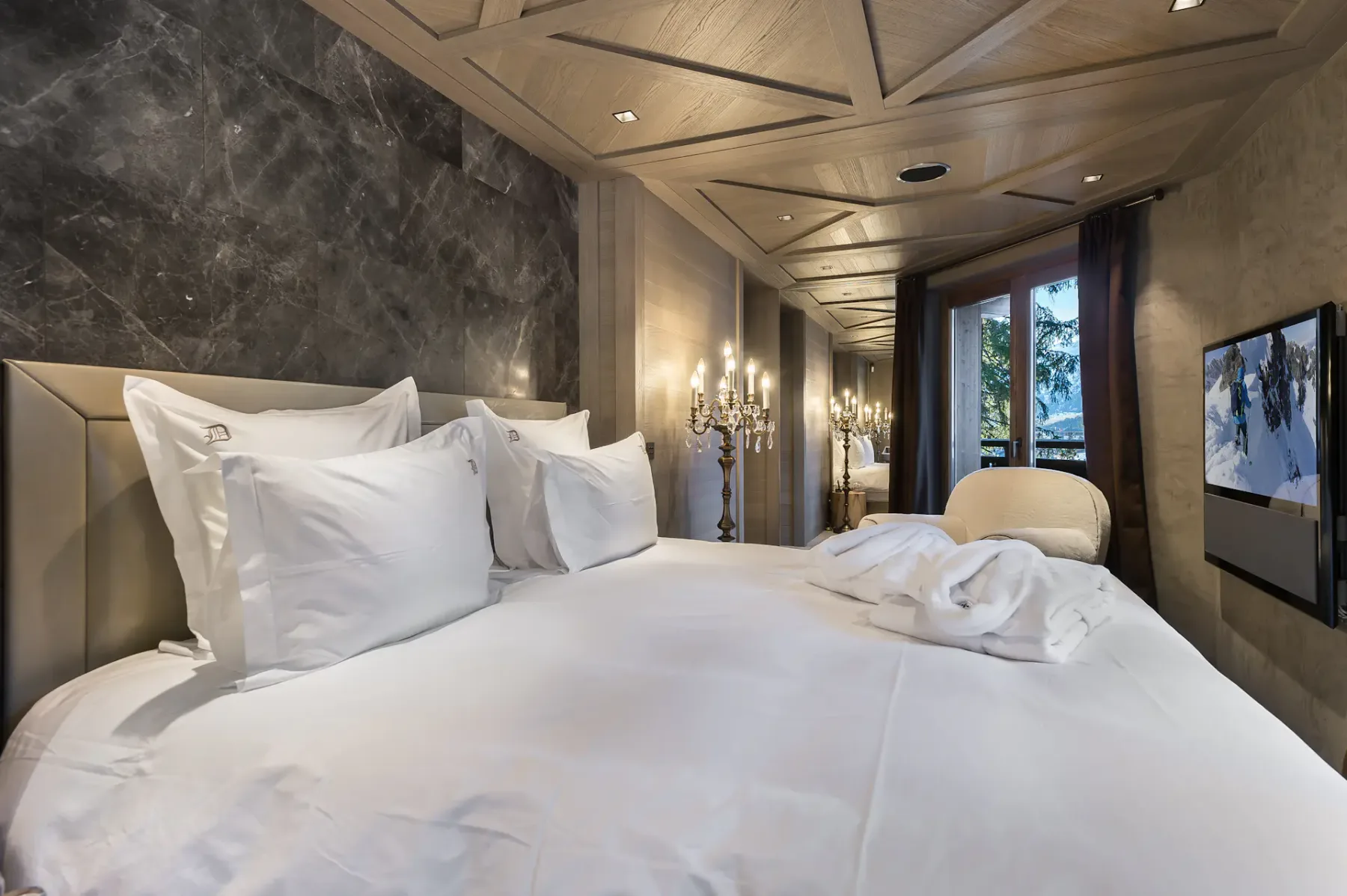 Lujosa habitación de hotel con una cama grande, ropa de cama blanca, una lámpara de araña y un televisor montado en la pared. Una ventana ofrece vistas a la vegetación exterior.