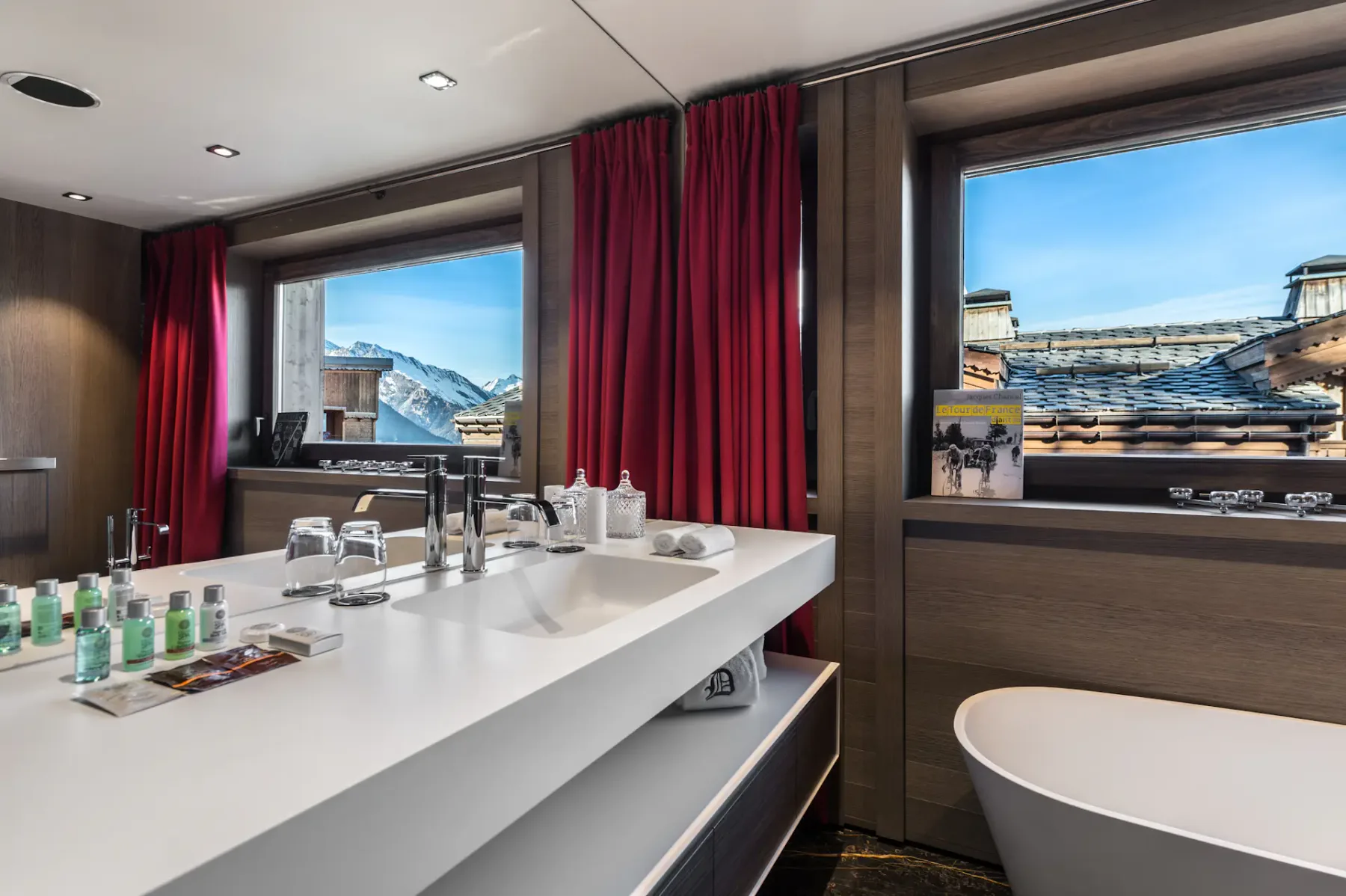 Baño moderno con una elegante encimera blanca, artículos de aseo, cortinas rojas, dos grandes ventanas con vistas a las montañas y una bañera exenta.