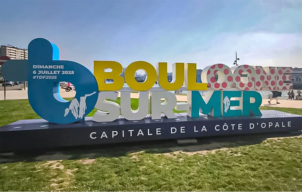 A large colorful event sign stands on a grassy outdoor plaza.