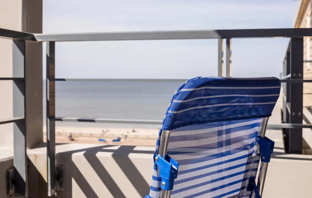 A balcony overlooks the beach, with a blue striped folding chair in the foreground.
