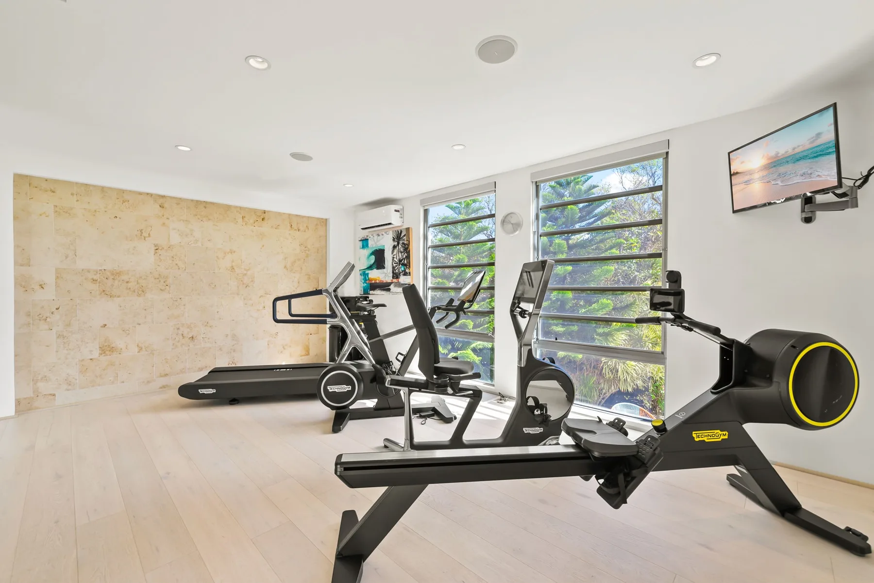 A bright exercise room with cardio equipment beside large windows.