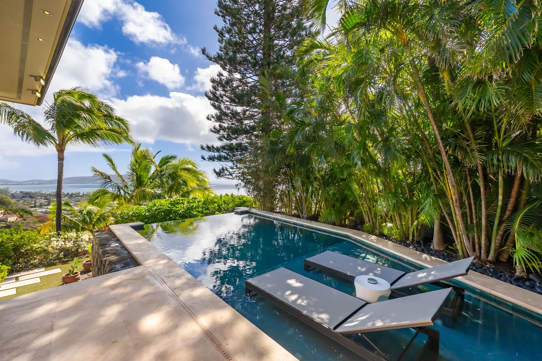 An outdoor swimming pool beside tropical plants, with lounge seating and a distant coastal view.